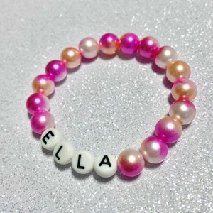 Puede incluir: Un brazalete de cuentas de color rosa y blanco degradado con el nombre "ELLA" escrito en cuentas de letras blancas.