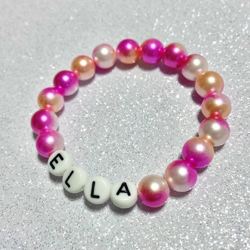 Puede incluir: Un brazalete de cuentas de color rosa y blanco degradado con el nombre "ELLA" escrito en cuentas de letras blancas.