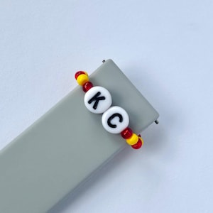 May include: A red and yellow beaded bracelet with white letter beads spelling "KC".