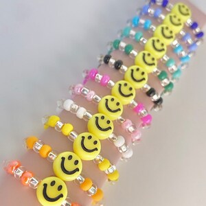 May include: A set of colorful beaded bracelets with yellow smiley face charms. The bracelets are made with small, round beads in various colors, including pink, white, orange, yellow, green, blue, and black. Each bracelet features a single yellow smiley face charm.