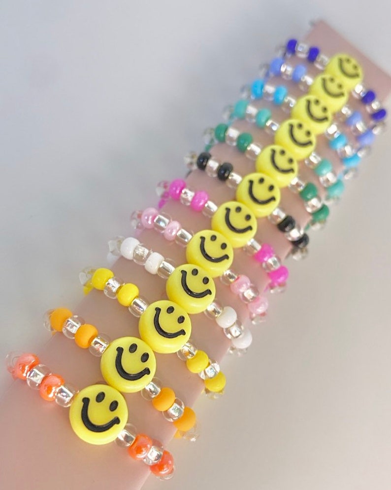 May include: A set of colorful beaded bracelets with yellow smiley face charms. The bracelets are made with small, round beads in various colors, including pink, white, orange, yellow, green, blue, and black. Each bracelet features a single yellow smiley face charm.