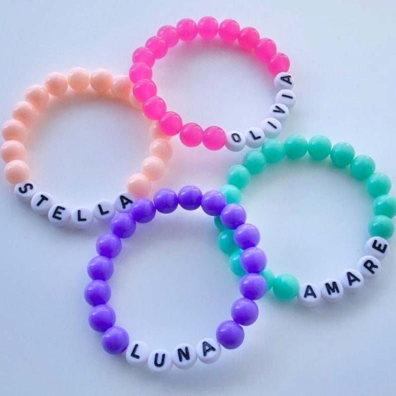 Puede incluir: Cuatro pulseras de cuentas de colores con cuentas de letras blancas que deletrean los nombres "Stella", "Olivia", "Luna" y "Amare".
