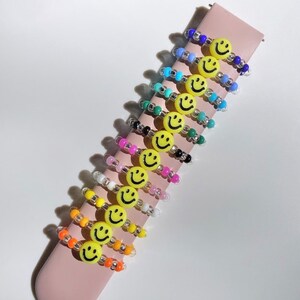 May include: A set of 10 colorful beaded rings with a yellow smiley face charm on each ring. The rings are stacked on a pink surface.