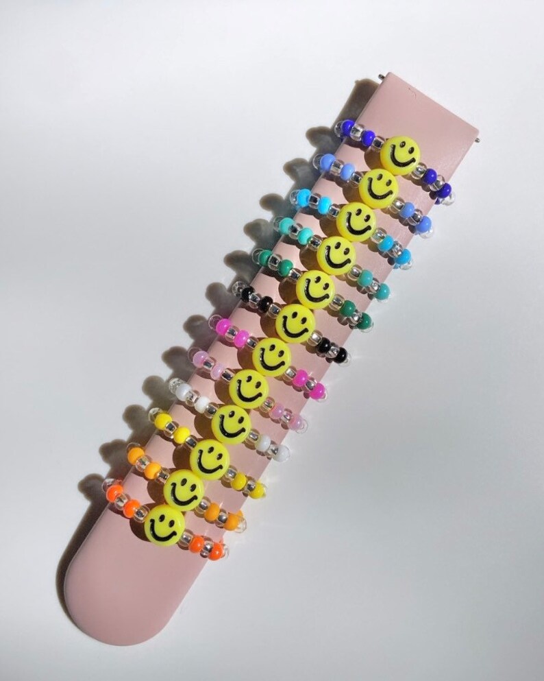 May include: A set of 10 colorful beaded rings with a yellow smiley face charm on each ring. The rings are stacked on a pink surface.