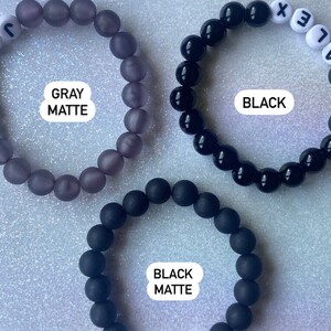 Puede incluir: Tres pulseras de cuentas, una gris mate, una negra mate y una negra brillante. Las pulseras tienen cuentas de letras blancas que deletrean "ALEX" y "JON".