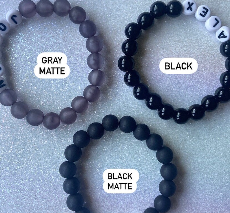 Puede incluir: Tres pulseras de cuentas, una gris mate, una negra mate y una negra brillante. Las pulseras tienen cuentas de letras blancas que deletrean "ALEX" y "JON".