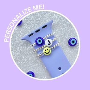 May include: A purple Apple Watch band with three charms: a blue evil eye, a white letter J, and a yellow smiley face.