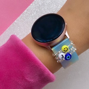 May include: A rose gold smartwatch with a black face and a blue and white patterned watch band. The watch band has a silver chain with charms, including a smiley face, a letter J, and a blue evil eye.