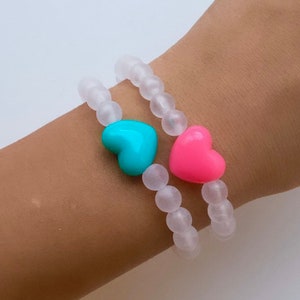 May include: Two bracelets with frosted white beads and heart-shaped charms. One charm is turquoise blue and the other is pink.
