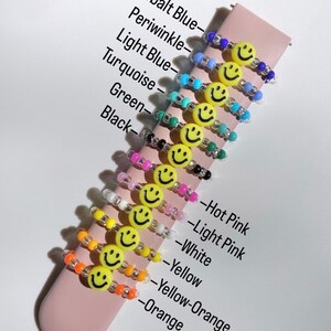 May include: A set of 12 colorful beaded bracelets with a smiley face charm. The bracelets are in the colors: cobalt blue, periwinkle, light blue, turquoise, green, black, hot pink, light pink, white, yellow, yellow-orange, and orange.