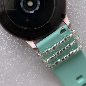 May include: A mint green smartwatch band with a silver beaded bracelet wrapped around it.