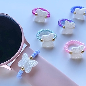 Può includere: Un set di sei anelli colorati con perline e ciondoli a forma di farfalla bianca. Gli anelli sono realizzati con piccole perline di vari colori, tra cui rosa, blu e viola. Ogni anello presenta un ciondolo a forma di farfalla bianca con dettagli dorati.