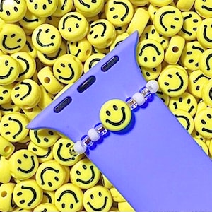 Può includere: Una custodia per telefono viola con un braccialetto di perline bianche e blu attaccato. Il braccialetto ha un ciondolo a forma di faccina sorridente gialla. La custodia del telefono è circondata da perline a forma di faccina sorridente gialle.