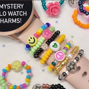 May include: Five mystery halo watch charms with different designs, including a smiley face, a star, a heart, a peace sign, and a star. The charms are made of colorful beads and are attached to a watch band.
