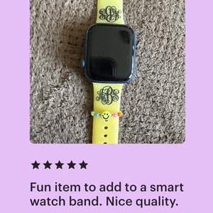May include: A yellow silicone watch band with a black monogram on each side. The band is on a smartwatch with a black face.
