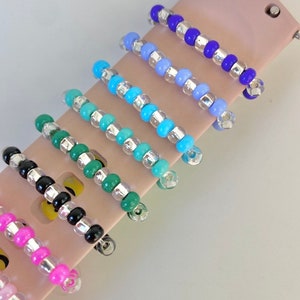 May include: Seven colorful beaded watch bands with silver accents. The bands are arranged in a row on a pink surface.