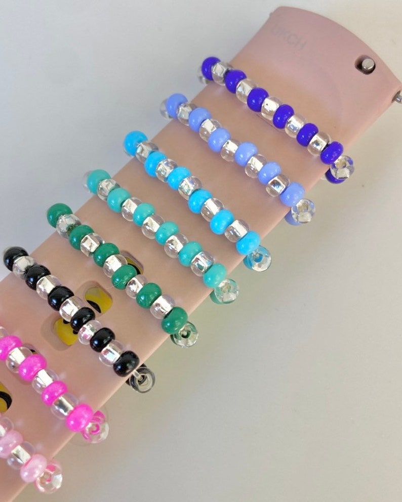 May include: Seven colorful beaded watch bands with silver accents. The bands are arranged in a row on a pink surface.