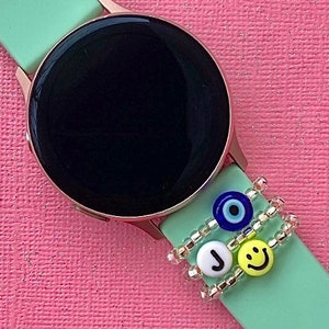 May include: A mint green smartwatch with a black screen and a gold bezel. The watch has a beaded bracelet with a blue evil eye charm, a white letter J charm, and a yellow smiley face charm.