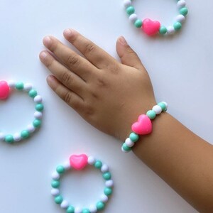 May include: Three bracelets made with white and light green beads with a pink heart charm. One bracelet is on a child's wrist.