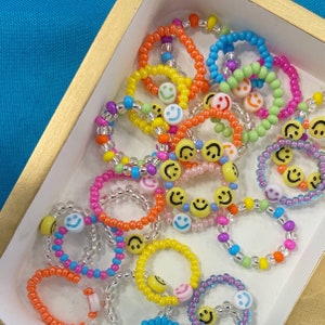 May include: A collection of colorful beaded rings with smiley face charms. The rings are made with various colors of beads, including pink, blue, green, yellow, and orange. The smiley face charms are white with black eyes and a black smile.