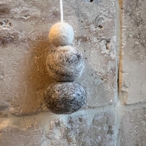 May include: A white, grey and black felt ball ornament hanging from a white string. The ornament is made of three felt balls stacked on top of each other.
