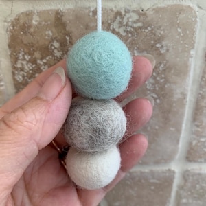 May include: A set of three felt balls in a light blue, grey, and white colour scheme. The balls are strung together and are hanging from a ceiling or wall.