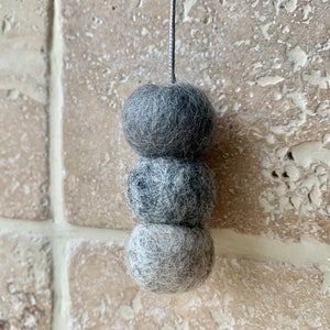May include: A set of three grey felt balls hanging from a string. The balls are different shades of grey and are arranged in a stack.