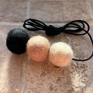 May include: Three felt balls in black, tan, and cream colours, connected by a black cord. The balls are round and have a textured, woolly appearance. The cord is looped and knotted.