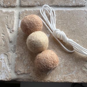 May include: Three felt balls in shades of beige and cream, arranged on a textured surface. A white rope is knotted and extends from the balls. The balls are likely part of a decorative or craft project.