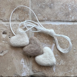 May include: Three heart-shaped felt ornaments in off-white and brown, strung on a white cord. The ornaments are displayed on a light-coloured stone surface. The cord is knotted at the top.