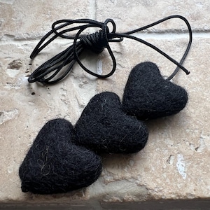 May include: Three black felt heart decorations with a black cord. The hearts are arranged in a row, and the cord is looped at the top. The decorations are on a light-coloured, textured surface.
