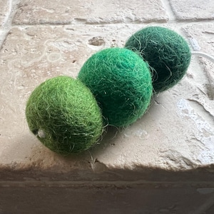 May include: Three green felt balls strung together to form a caterpillar. The balls are a gradient of green, with the first ball being the lightest green and the last ball being the darkest green.