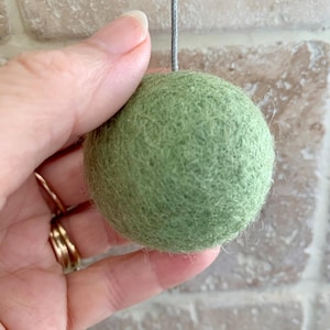 May include: A green felt ball with a string attached to the top. The ball is being held in a hand.