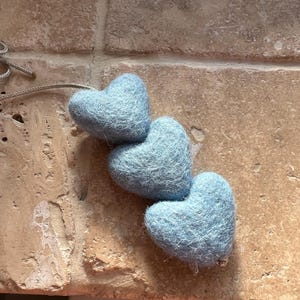 Könnte beinhalten: Eine Kette aus drei hellblauen Filzherzen, die durch eine dünne, hellfarbige Schnur verbunden sind. Die Herzen sind diagonal auf einer strukturierten, hellbraunen Oberfläche angeordnet. Der Filz hat eine weiche, flauschige Textur.