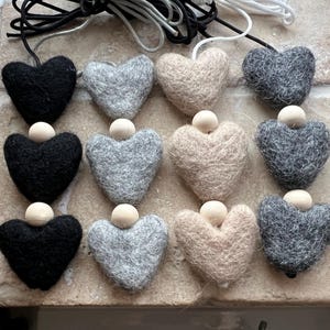 Puede incluir: Un conjunto de adornos de fieltro en forma de corazón en negro, gris y beige, cada uno con una pequeña cuenta de madera en la parte superior. Los adornos están ensartados en cordones blancos y negros, para decorar.