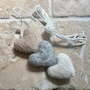 May include: Three felt heart decorations in beige, grey, and white, joined by a white string. The hearts have a soft, textured appearance, with a simple, rustic design. The string is knotted at the top, ready for hanging.