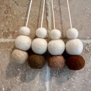May include: Close-up of four decorative felt ball ornaments. Each ornament has three felt balls in varying sizes and colours: white, tan, brown, and dark brown. The ornaments hang from white cords against a neutral background.