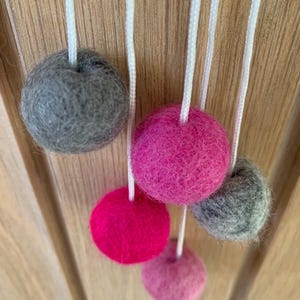 May include: A garland of felt balls in shades of pink and grey, hanging from white strings. The balls are arranged in a staggered pattern, with the pink balls at the top and the grey balls at the bottom.
