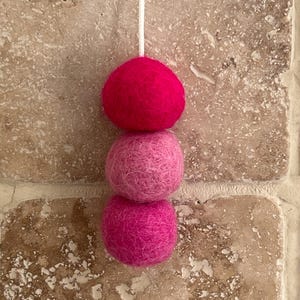 May include: Three pink felt balls hanging from a white string. The balls are different shades of pink, with the top ball being the darkest and the bottom ball being the lightest.