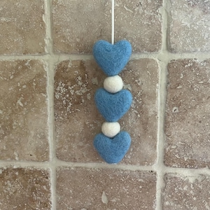 Puede incluir: Adorno colgante con tres corazones de fieltro azul claro y dos bolas de fieltro blanco. Los corazones están espaciados uniformemente y conectados por una cuerda blanca, creando un diseño encantador. El fondo es una superficie texturizada de tonos neutros.