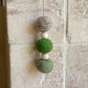 May include: A hanging mobile with three felt balls and wooden beads. The top ball is light brown, the middle ball is green, and the bottom ball is a darker shade of green. The wooden beads are natural wood colour.