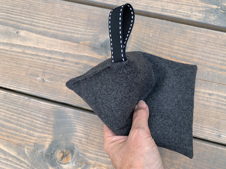 May include: A gray felt doorstop with a black fabric handle. The handle has white stitching.