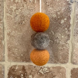 May include: Three felt balls in shades of orange and grey suspended by a white string. The top and bottom balls are orange, while the middle ball is grey. The balls are hanging against a brick wall.