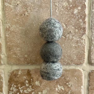 May include: A string of three grey felt balls hanging from a white string. The balls are different sizes and are all made of a soft, fuzzy material.