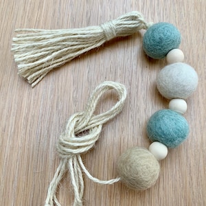 May include: A garland of felt balls in shades of green and beige, strung with twine. The garland has a tassel at one end.
