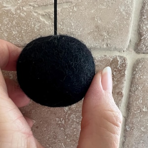 May include: A black felt ball, suspended by a black string, is held between fingers. The ball has a textured surface and is round. The background is a light brown brick wall.