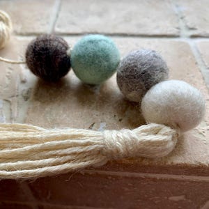 May include: A garland of felt balls in brown, green, grey, and white colours with a natural fibre tassel.
