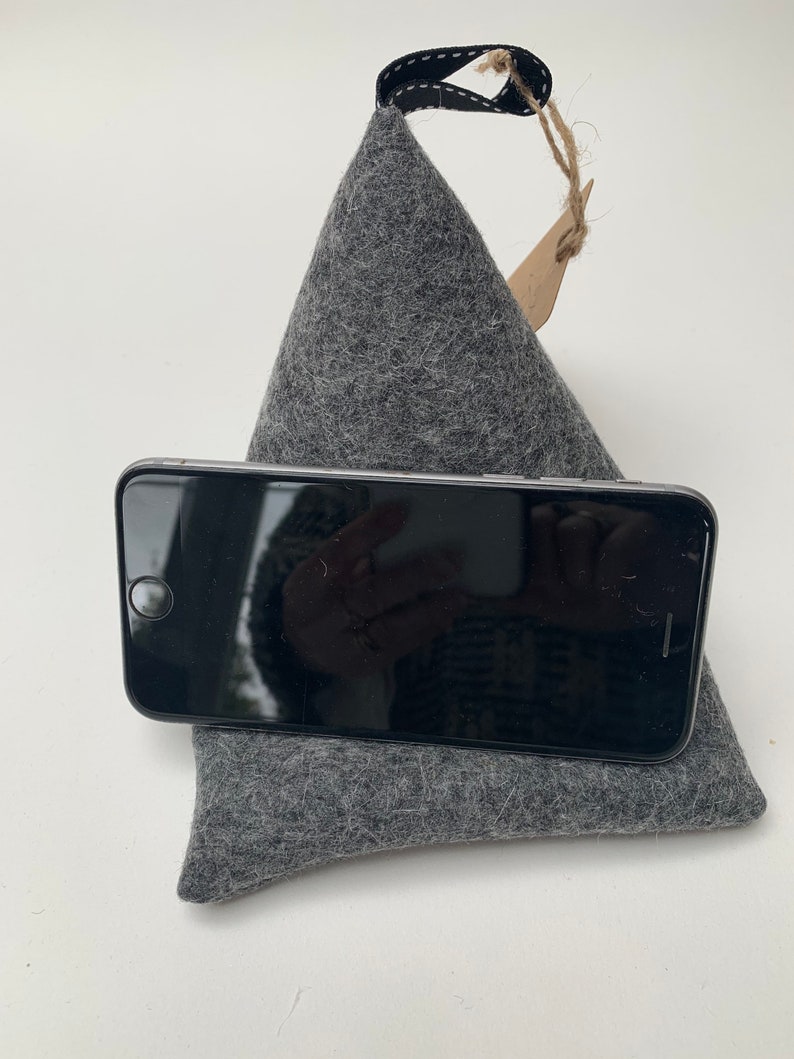 May include: A gray felt phone stand with a black loop handle. The stand is shaped like a pyramid and has a small brown tag attached to the handle.