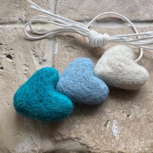 May include: Three felt heart ornaments in teal, light blue, and white, connected by a white string. The hearts are arranged in a row, with the string looped above them. The ornaments are on a textured, light brown surface.