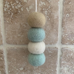 May include: A string of four felt balls in shades of beige, light blue, white, and light blue. The balls are strung together and hanging from a white cord.